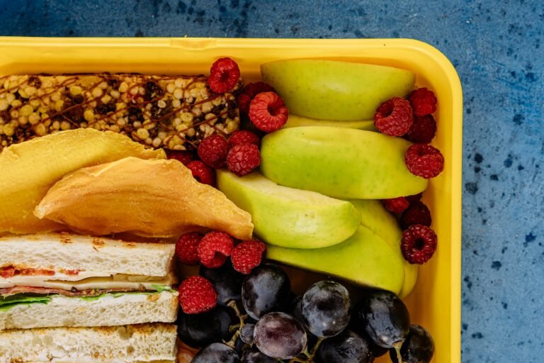 Colorful lunchbox containing apples, grapes, raspberries, sandwich and cereal bar.